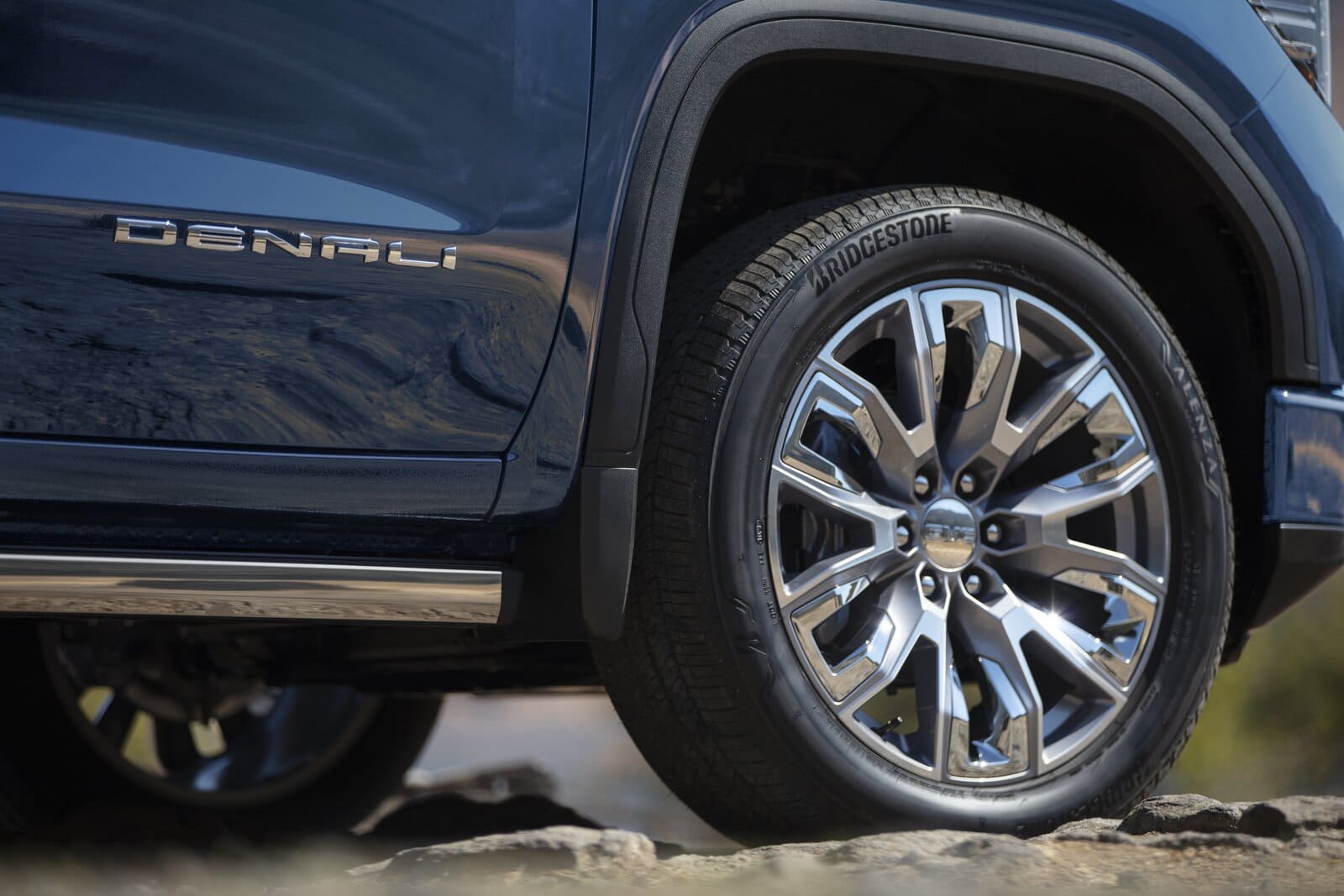 2025 GMC Sierra 1500 Denali - Felge Das Bild zeigt eine Detailaufnahme der Heckseite eines GMC Sierra 1500 Denali, bei der der Fokus auf der edlen, glanzgedrehten Mehrspeichen-Leichtmetallfelge und dem darauf montierten Bridgestone-Reifen liegt. Direkt über dem Schweller ist das "DENALI"-Emblem in Chrom angebracht, was den luxuriösen Charakter und die exklusive Modelllinie des Fahrzeugs unterstreicht. Der Truck steht auf unebenem Gelände, was die Verbindung von Eleganz und Geländetauglichkeit betont.