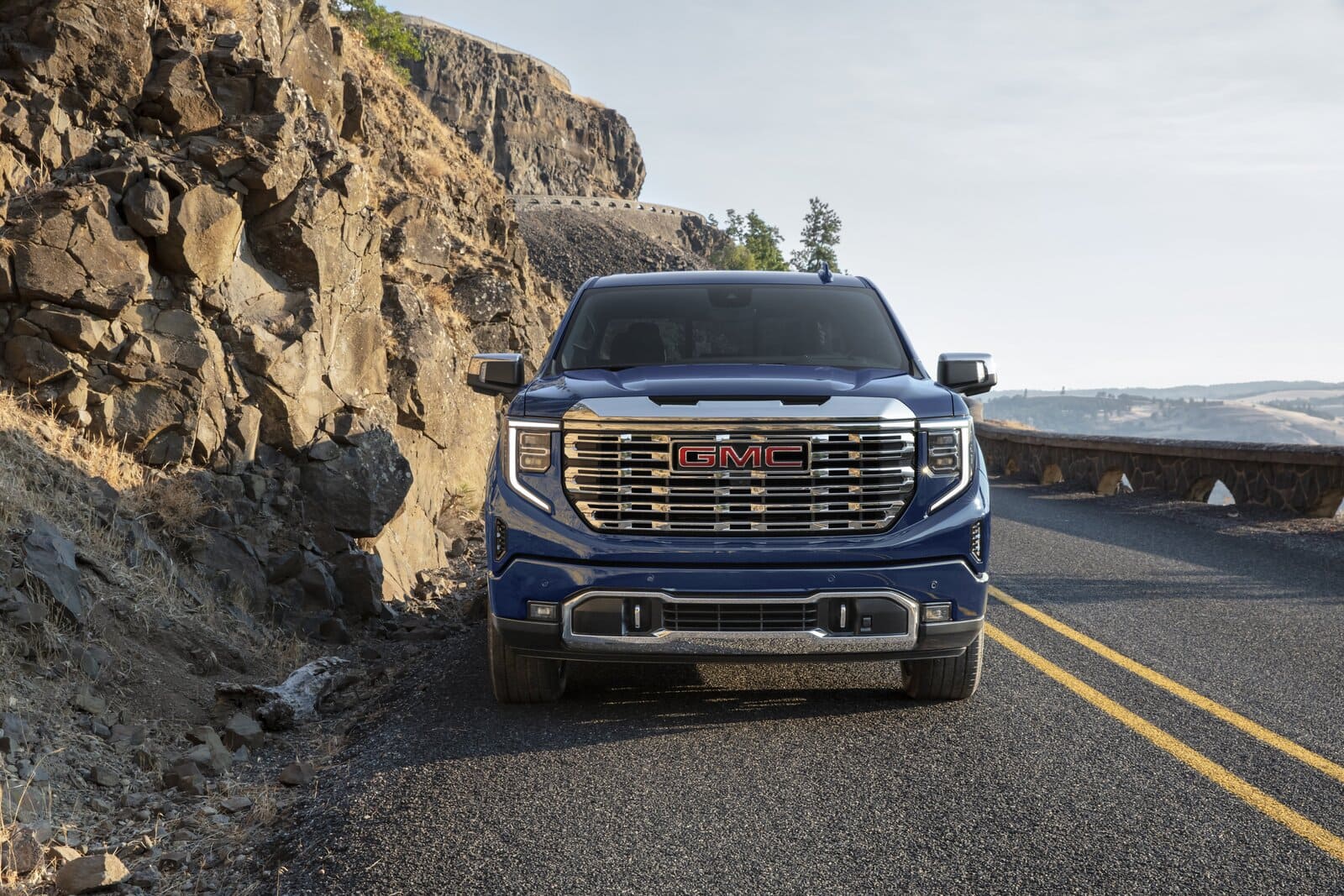 2025 GMC Sierra 1500 Denali - Frontansicht Das Bild zeigt den 2025 GMC Sierra 1500 Denali aus der Frontalperspektive, wie er kraftvoll auf einer kurvenreichen Bergstraße steht. Dominant präsentiert sich der großflächige, hochglänzende Denali-Kühlergrill mit rotem GMC-Logo, flankiert von vertikal angeordneten LED-Scheinwerfern. Die felsige Landschaft auf der linken Seite und der weite Blick ins Tal auf der rechten Seite unterstreichen den souveränen, luxuriösen und zugleich abenteuerbereiten Auftritt des Fahrzeugs.