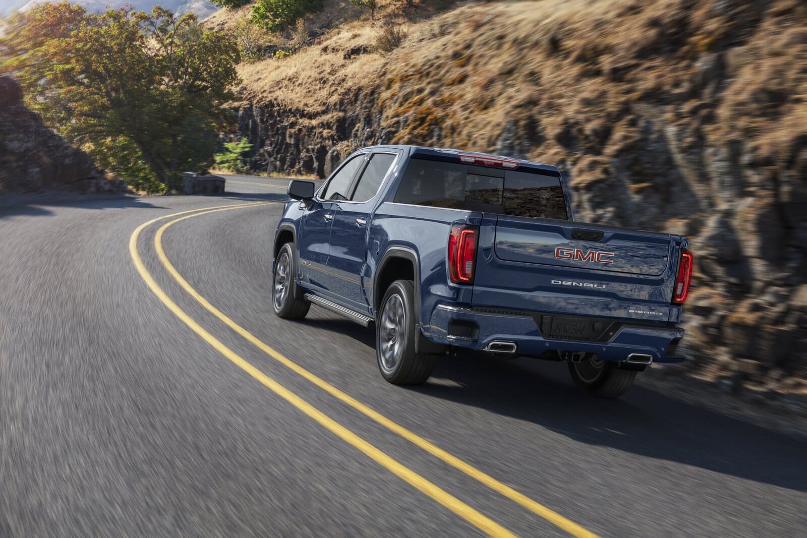 2025 GMC Sierra 1500 Denali - Heckansicht Das Bild zeigt den 2025 GMC Sierra 1500 Denali in der Heckansicht, wie er dynamisch durch eine kurvige Bergstraße fährt. Der dunkelblaue Truck präsentiert sich mit der charakteristischen "DENALI"-Prägung auf der Heckklappe, GMC-Logo in Rot, vertikalen LED-Rückleuchten und doppelflutiger Abgasanlage. Die Szene vermittelt Luxus und Leistungsfähigkeit in einer landschaftlich reizvollen, naturverbundenen Umgebung.