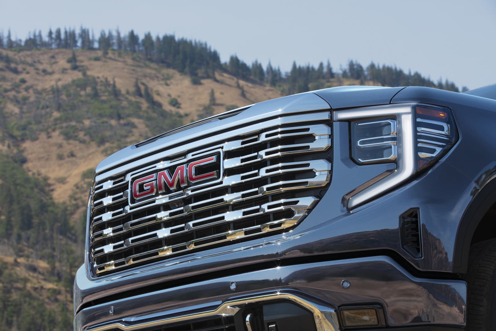 2025 GMC Sierra 1500 Denali - Kühlergrill Das Bild zeigt eine eindrucksvolle Nahaufnahme der Front des 2025 GMC Sierra 1500 Denali in einer natürlichen Outdoor-Kulisse mit bewaldeten Berghängen im Hintergrund. Der massive, glänzend verchromte Kühlergrill mit integriertem roten GMC-Logo dominiert das Bild und wird flankiert von modernen, vertikal verlaufenden LED-Tagfahrlichtern – ein Design, das Stärke, Luxus und technologische Raffinesse perfekt vereint.