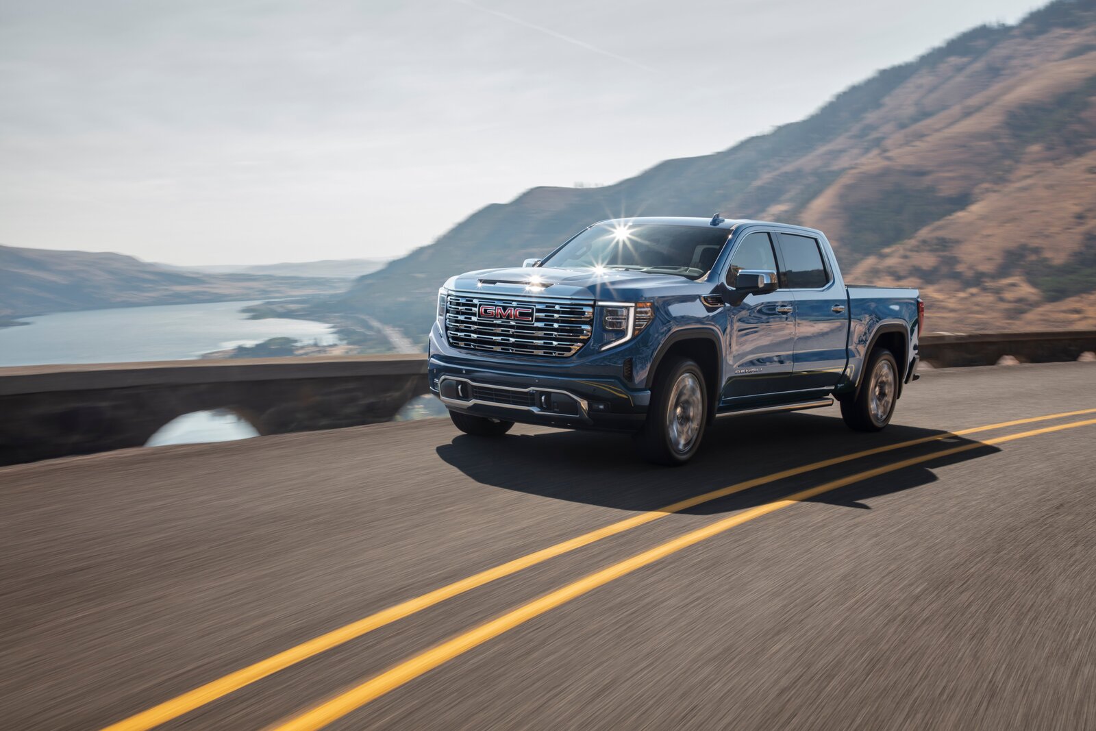 2025 GMC Sierra 1500 Denali - Seitenansicht as Bild zeigt den 2025 GMC Sierra 1500 Denali in Bewegung auf einer kurvenreichen Panoramastraße mit Blick auf ein malerisches Flusstal und bewaldete Berghänge. Der luxuriöse Pickup glänzt im Sonnenlicht, wobei insbesondere der großflächige verchromte Kühlergrill mit dem roten GMC-Logo und die markanten LED-Tagfahrlichter ins Auge fallen. Die Szenerie unterstreicht den Premium-Charakter des Fahrzeugs ebenso wie seine Fähigkeit, Komfort und Leistungsstärke mit Abenteuerlust zu vereinen.