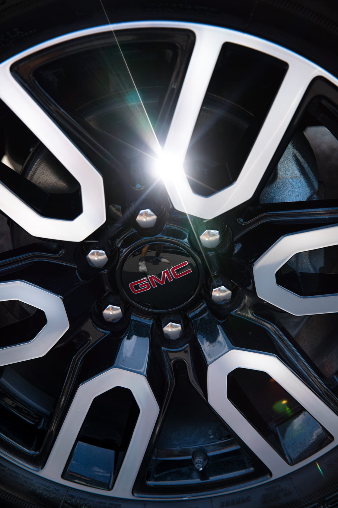 GMC Sierra 1500 AT4 Felge Das Bild zeigt eine Nahaufnahme einer glänzenden, schwarz-silbernen Leichtmetallfelge des GMC Sierra 1500 AT4. In der Mitte prangt das rot-silberne GMC-Logo, während die Sonnenreflexion auf dem Metall die hochwertige Verarbeitung und das markante Design der Felge betont.