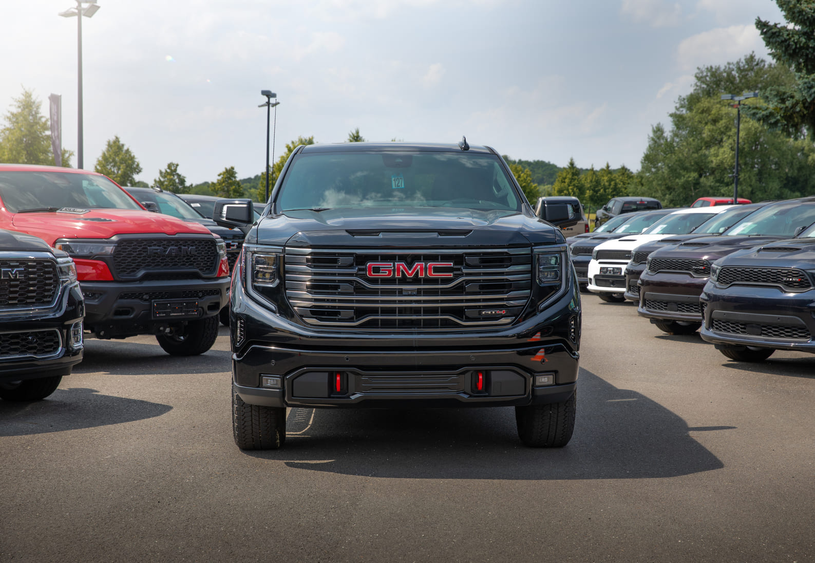 GMC Sierra 1500 AT4 Frontansicht Das Bild zeigt die eindrucksvolle Frontansicht eines GMC Sierra 1500 AT4, der mittig in einer Reihe von US-Trucks auf einem Autohaus-Gelände steht. Der schwarze Truck mit dominantem GMC-Logo, breitem Kühlergrill und roten Tow Hooks vermittelt Kraft und Präsenz – perfekt eingerahmt von weiteren RAM- und Dodge-Modellen im Hintergrund.