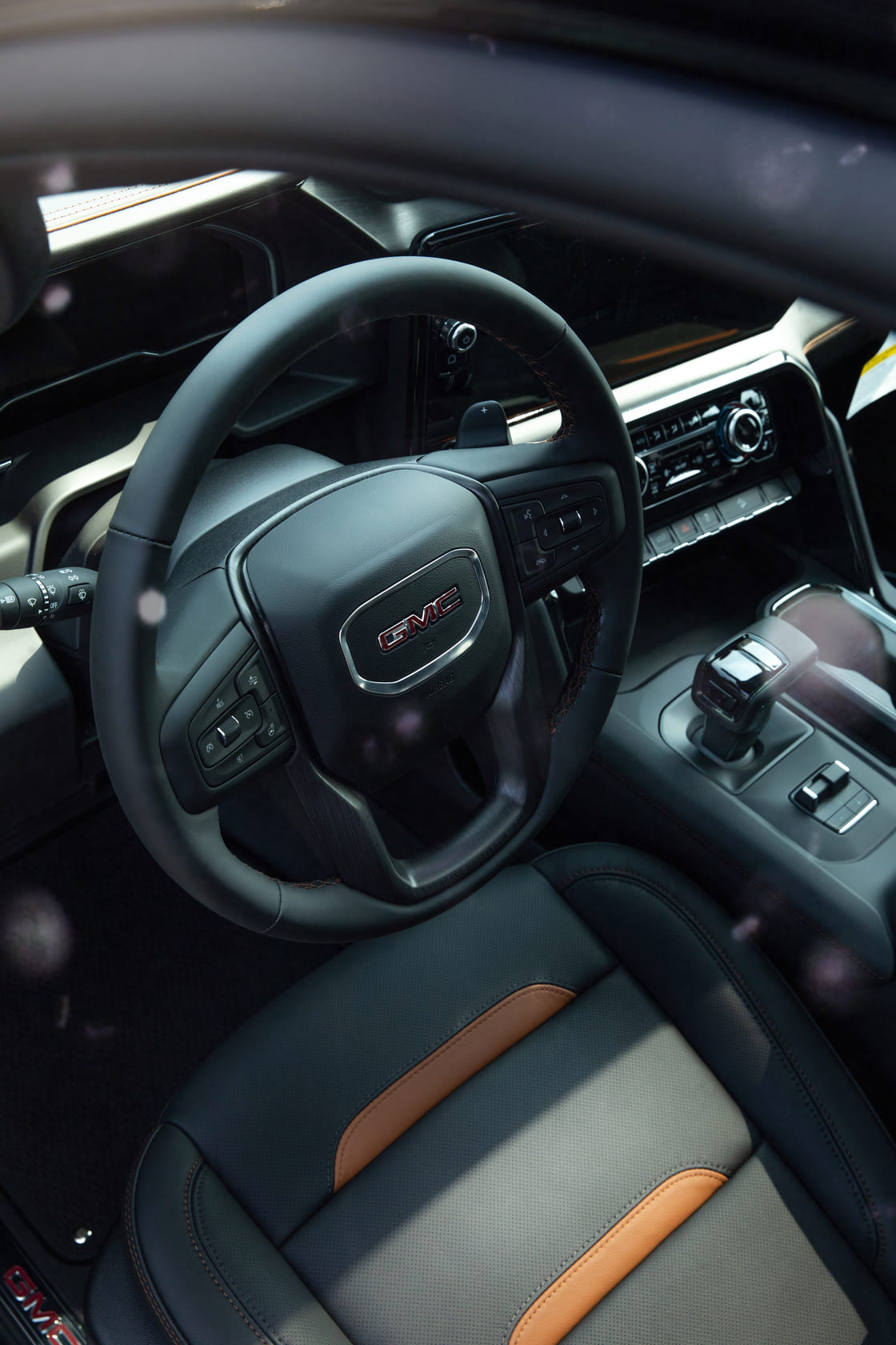Das Bild zeigt den Innenraum eines GMC Sierra 1500 AT4 mit edler Ausstattung: ein lederbezogenes Multifunktionslenkrad mit GMC-Logo, zentraler Schalthebel und moderne Bedienelemente. Die perforierten Ledersitze in Schwarz mit braunen Kalahari-Akzenten unterstreichen den hochwertigen, zugleich robusten Charakter des Trucks.