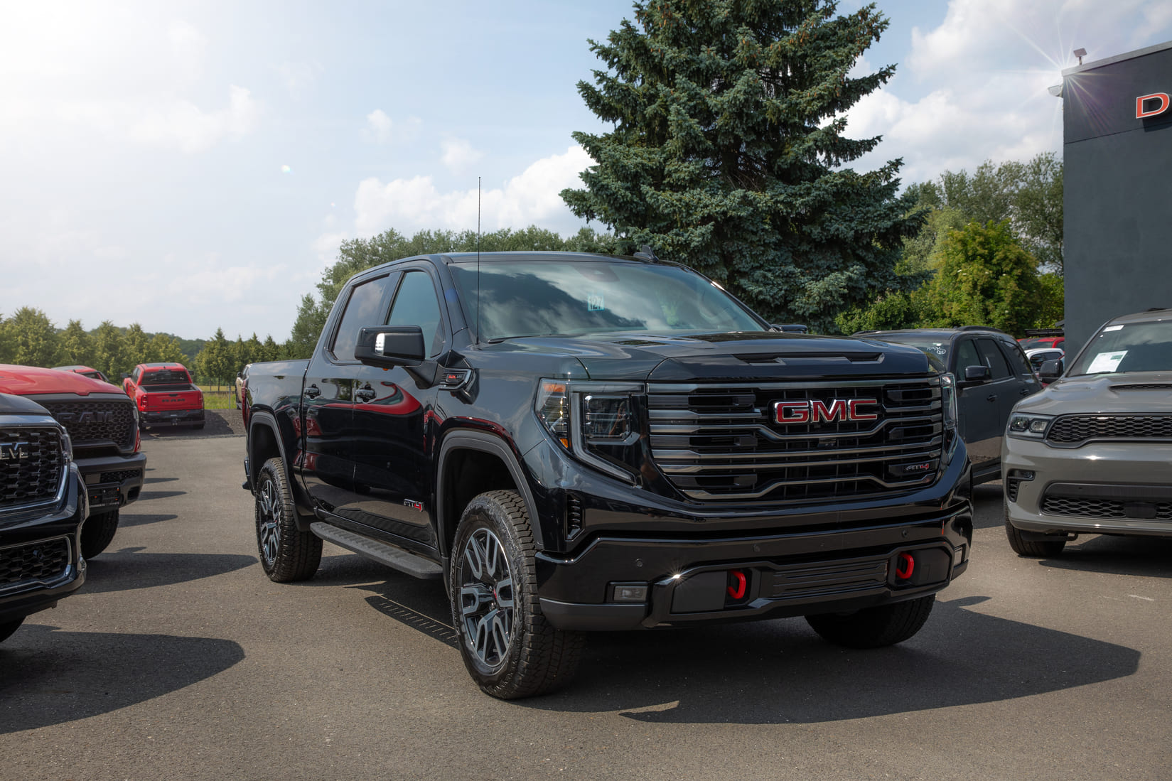 Auf dem Bild ist ein schwarzer GMC Sierra 1500 AT4 zu sehen, der prominent auf dem Hof eines US-Fahrzeughändlers steht. Das markante Design mit schwarzem Kühlergrill, roten Tow Hooks und AT4-Logo unterstreicht den Offroad-Charakter des Trucks. Im Hintergrund sind weitere US-Pickups und ein Verkaufsgebäude zu erkennen – die Szene vermittelt echte American-Car-Atmosphäre.